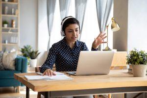 Woman in a blue shirt working from home in her office, wearing headphones.